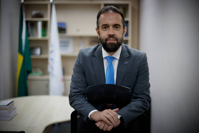 Um homem com barba e cabelo curto está sentado em uma cadeira, com as mãos cruzadas sobre o colo. Ele usa um terno escuro e uma gravata azul. Ao fundo, há uma mesa de escritório e prateleiras com livros e documentos. Uma bandeira do Brasil está visível à esquerda.
