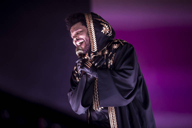 O cantor The Weeknd está se apresentando em um palco, usando um manto preto com detalhes dourados e uma capuz. Ele está segurando um microfone e sorrindo enquanto canta. O fundo é iluminado em tons de roxo, criando uma atmosfera vibrante.