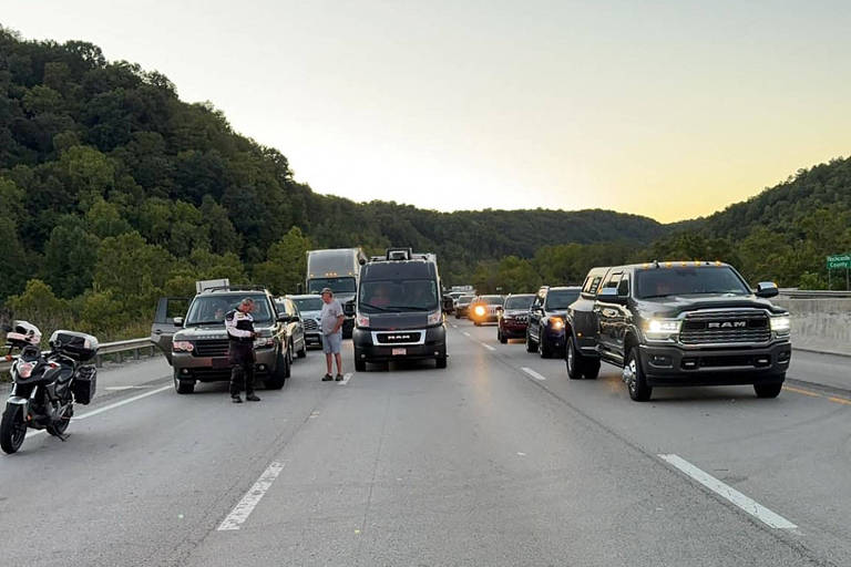 Foto mostra alguns veículos estacionados em uma rodovia de três faixas. Alguns motoristas desembarcaram do carro