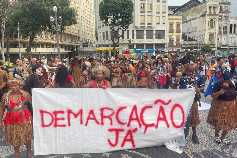 A imagem mostra um grupo de pessoas participando de um protesto em uma área urbana. Muitas delas estão vestidas com trajes tradicionais indígenas, algumas usando penas e adornos. Um grande banner branco com a inscrição 'DEMARCAÇÃO JÁ' em letras vermelhas é segurado por alguns manifestantes. O ambiente é urbano, com prédios ao fundo e outras pessoas observando o protesto.