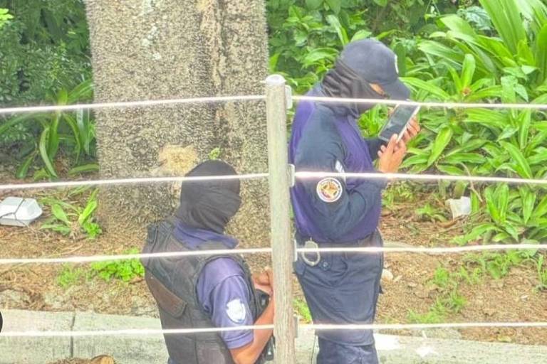 Imagem mostra dois homens fardados, encapuzados, paradas em uma rua. Pela foto é possível ver que já um muro com cerca de proteção em sua extremidade 