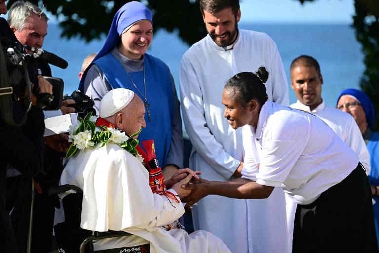 O papa Francisco, sentado em uma cadeira de rodas e usando vestes brancas, é saudado por uma mulher que se aproxima para cumprimentá-lo. Ao fundo, há várias pessoas, incluindo um homem e uma mulher com roupas religiosas, além de uma mulher usando um hábito. O cenário é ao ar livre, com árvores e o mar visível ao fundo.