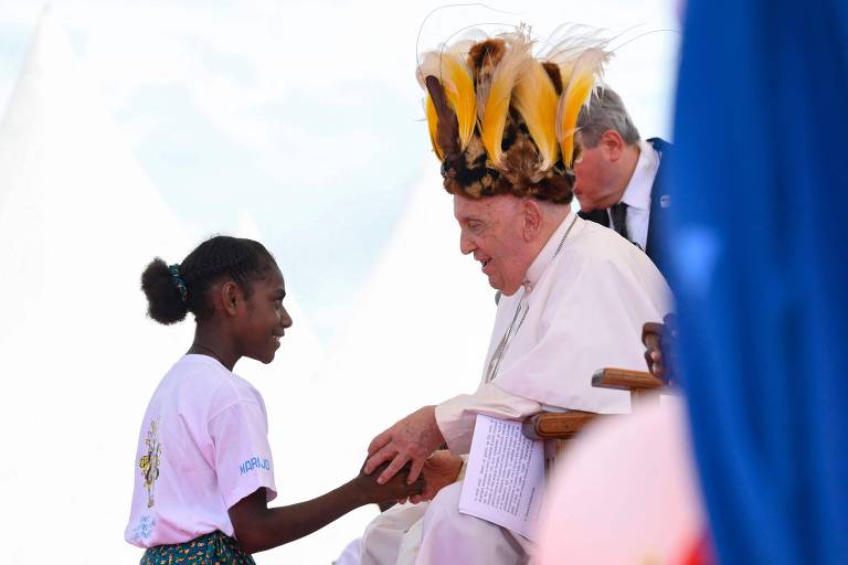 A imagem mostra uma jovem de pele escura, vestindo uma camiseta branca e uma saia colorida, cumprimentando o Papa Francisco, que está usando um chapéu decorado com penas. O Papa está sentado em uma cadeira, sorrindo e estendendo a mão para a jovem. Ao fundo, há uma pessoa de terno e um cenário com tendas brancas.