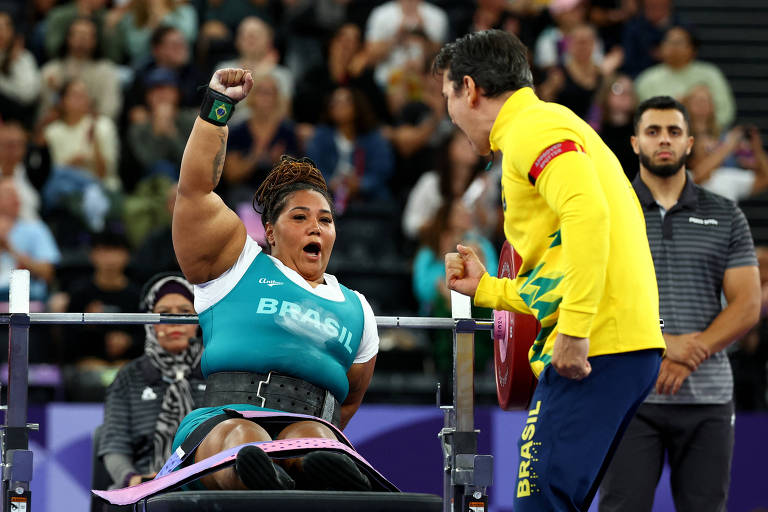 A imagem mostra uma atleta paralímpica comemorando com entusiasmo, levantando o braço, enquanto um treinador, vestido com uma jaqueta amarela com detalhes verdes (representando a equipe brasileira), reage de maneira energética em frente a ela. A atleta quebrou recorde na paralímípica de Paris no levantamento de peso adaptado. No fundo, o público acompanha o momento.