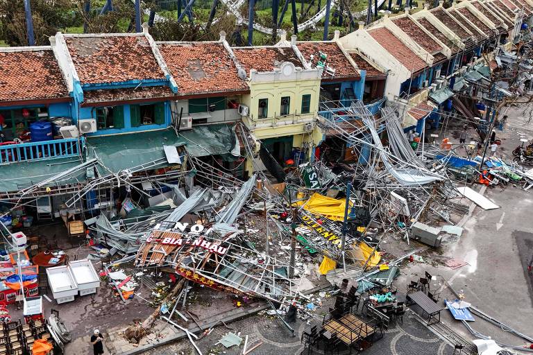 A imagem mostra uma área urbana devastada, com prédios danificados e estruturas metálicas caídas. As fachadas dos edifícios estão desmoronadas, e há destroços espalhados pelo chão. Algumas pessoas podem ser vistas na cena, e há sinais de destruição significativa, como fios elétricos expostos e objetos quebrados.