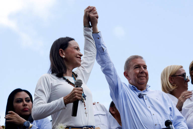 A imagem mostra duas pessoas em um evento político, levantando as mãos juntas em sinal de união. A mulher à esquerda, vestindo uma blusa branca e segurando um microfone, sorri enquanto levanta a mão. O homem à direita, com uma camisa azul clara, também levanta a mão. Ao fundo, outras pessoas estão presentes, algumas olhando para a frente e outras em diferentes poses.