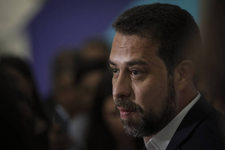 Em primeiro plano, com o fundo desfocado em que aparece um grupo de pessoas, Boulos, um homem branco, jovem, barba e cabelo castanhos, usando blazer preto e camisa branca. O rosto dele aparece de perfil, com o semblante sério