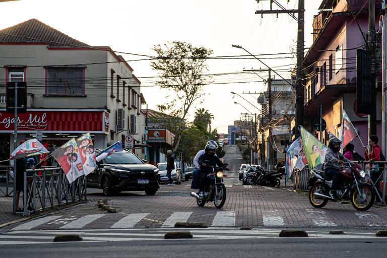 A imagem mostra um cruzamento urbano ao entardecer, com uma rua pavimentada e várias bandeiras coloridas expostas. Um carro preto está parado, enquanto duas motos estão em movimento, uma delas atravessando a faixa de pedestres. No fundo, é possível ver edifícios e postes de luz, além de algumas pessoas caminhando.