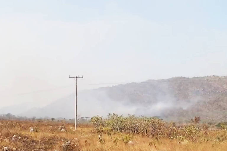 A imagem mostra uma área de vegetação seca com fumaça visível ao fundo, indicando uma queimada. Há um poste de eletricidade à esquerda e uma colina ao fundo, onde a fumaça se dispersa no ar. O céu está claro, mas a presença da fumaça sinaliza um incêndio nas proximidades.