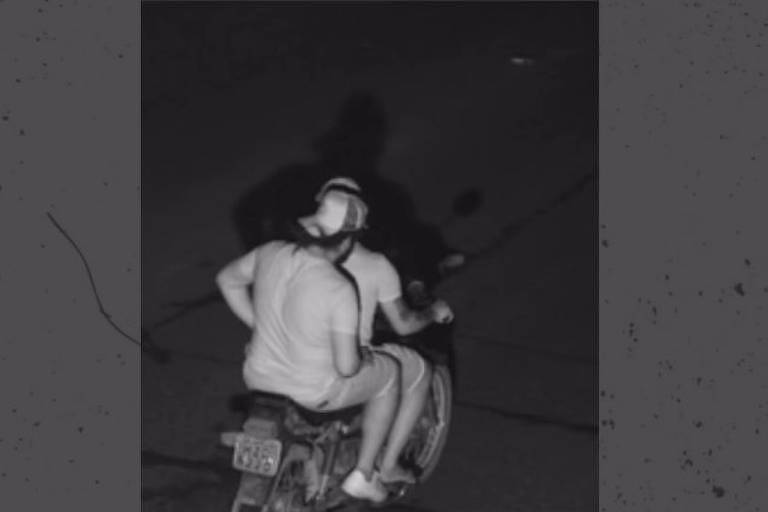 A imagem mostra duas pessoas em uma motocicleta, visíveis de costas. A cena é noturna, com pouca iluminação, e as sombras das pessoas são projetadas no chão. A pessoa da frente está usando uma camiseta e um boné, enquanto a pessoa de trás está vestindo uma camiseta de manga curta e shorts.