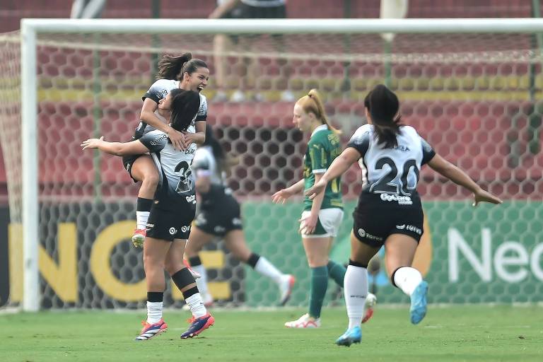 A imagem mostra duas jogadoras de futebol comemorando um gol. Uma jogadora está sendo levantada por outra, ambas vestindo uniformes brancos e pretos. Ao fundo, outras jogadoras estão em movimento, com uma delas vestindo um uniforme verde. O campo de futebol e a rede do gol são visíveis.
