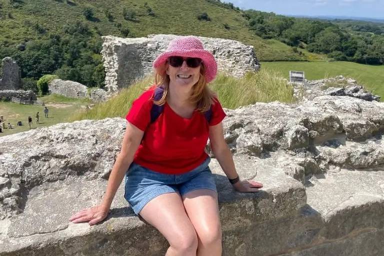 Uma mulher sentada em uma parede de pedra de ruínas, usando uma camiseta vermelha e um chapéu rosa. Ela está sorrindo e parece estar aproveitando o dia ensolarado. Ao fundo, há uma paisagem verde com colinas e algumas estruturas de pedra visíveis.