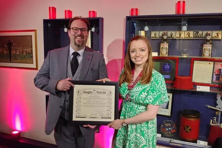 Duas pessoas estão posando para a foto, segurando um certificado. O homem à esquerda está vestido com um terno cinza e gravata preta, enquanto a mulher à direita usa um vestido verde. Ao fundo, há uma prateleira com objetos decorativos e iluminação em tons de rosa.