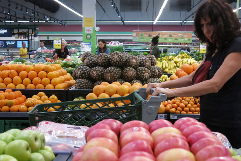 A imagem mostra uma seção de frutas em um supermercado. No primeiro plano, há uma variedade de maçãs vermelhas e verdes. Uma mulher com cabelo escuro e encaracolado está empurrando um carrinho de compras, observando as frutas. Ao fundo, há pilhas de laranjas, abacaxis e bananas, além de outras pessoas fazendo compras.
