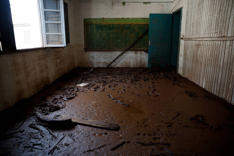 A imagem mostra uma sala abandonada com o chão coberto de lama. Há uma lousa verde na parede e uma porta azul aberta. A luz entra pela janela， iluminando o ambiente sujo e desolado.
