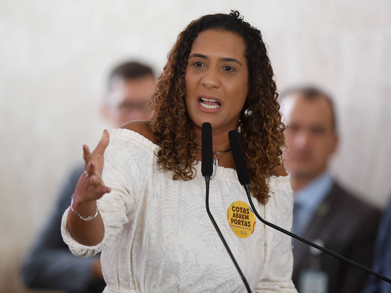 Uma mulher com cabelo cacheado e solto， vestindo uma blusa branca， está falando em um microfone. Ela gesticula com a mão direita enquanto expressa suas ideias. Ao seu lado， há homens em trajes formais， e ao fundo， uma parede clara. A mulher usa um adesivo amarelo com a frase 039;COTAS ABREM PORTAS039;.