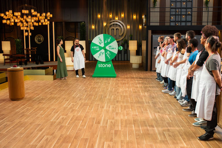  A imagem mostra um cenário de competição de culinária, com um grupo de participantes alinhados à direita, todos usando aventais brancos