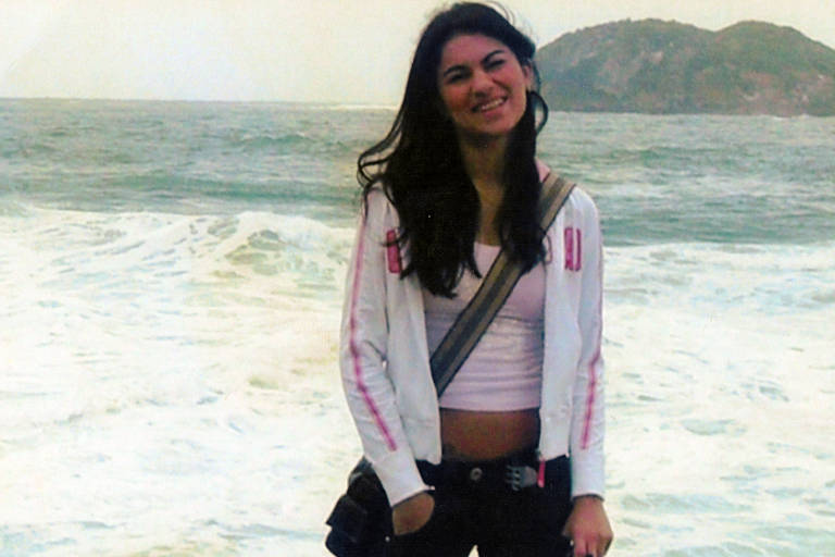 Uma jovem está sorrindo em uma praia, com ondas do mar ao fundo. Ela usa uma blusa de manga longa branca com detalhes em rosa e uma camiseta rosa por baixo. A jovem tem cabelo longo e solto, e está usando calças escuras. Ela segura uma bolsa preta e parece estar em um dia nublado