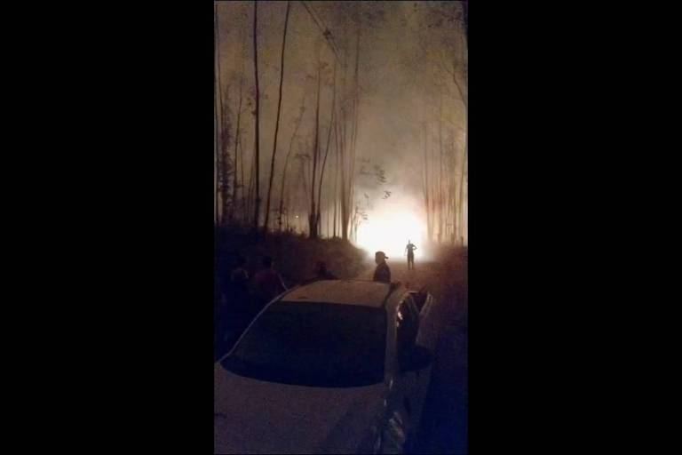 A imagem mostra um incêndio florestal em uma área com árvores altas. Há fumaça densa no ar, e uma luz intensa proveniente do fogo é visível ao fundo. Algumas pessoas estão paradas em uma estrada, observando a cena, enquanto um carro branco está estacionado à frente. A atmosfera é de tensão e preocupação devido à situação do incêndio.