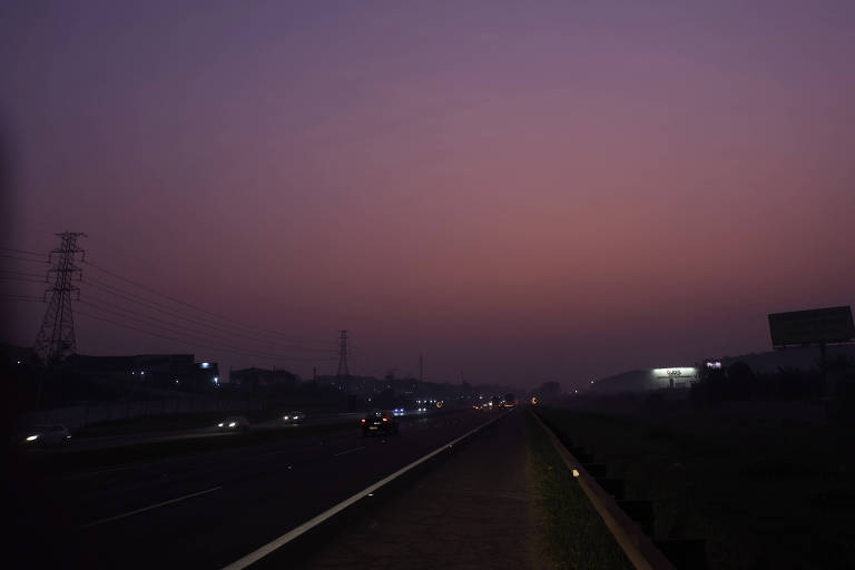 A imagem mostra uma estrada em um ambiente urbano ao amanhecer, com um céu em tons de roxo e azul. Há veículos se movendo na estrada, e à esquerda, pode-se ver postes de energia elétrica. O cenário é levemente iluminado pelas luzes dos carros e algumas construções ao fundo.