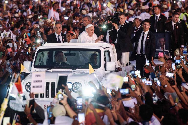 A imagem mostra o Papa em um carro aberto, acenando para uma multidão de pessoas que o cercam. Ele está cercado por seguranças e há várias pessoas segurando celulares e bandeiras. O ambiente é festivo, com muitas pessoas visíveis na multidão, algumas segurando cartazes e outras com expressões de alegria.