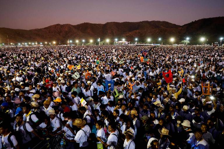 A imagem mostra uma grande multidão reunida em um evento ao ar livre, com pessoas vestindo camisetas brancas e chapéus. O cenário é montanhoso ao fundo, e a iluminação é suave, indicando que é ao entardecer. Algumas pessoas estão segurando bandeiras ou cartazes, e a multidão parece animada e engajada.