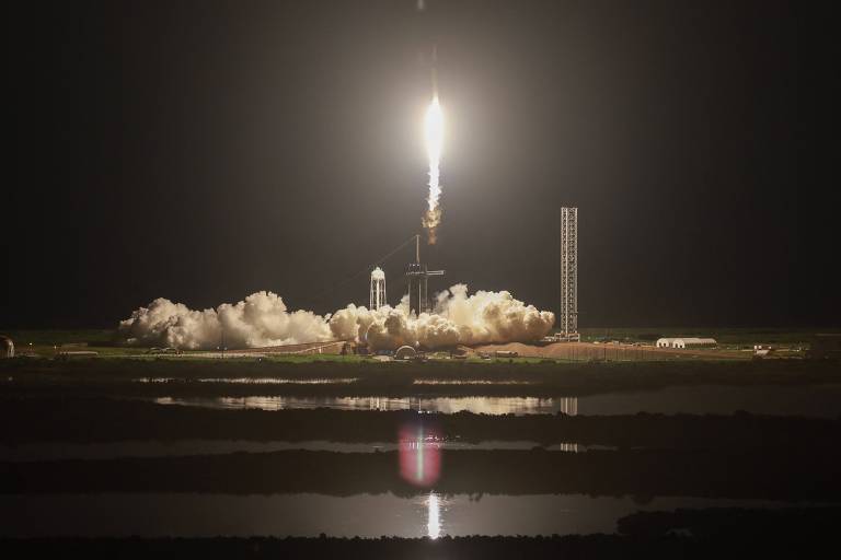 A imagem mostra um foguete sendo lançado à noite, com uma chama brilhante na parte inferior. A base do foguete está cercada por uma nuvem de fumaça e vapor, refletindo a luz do lançamento. O fundo é escuro, destacando a iluminação do foguete.
