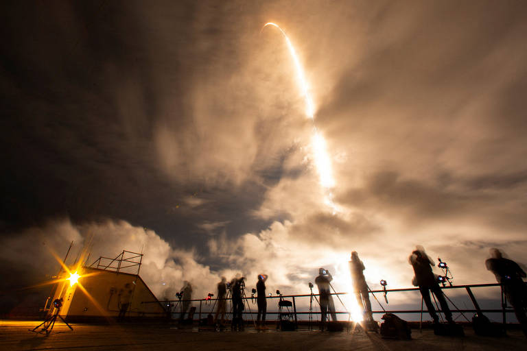 A imagem mostra um foguete sendo lançado ao céu, com uma trilha de luz brilhante. Silhuetas de várias pessoas estão em primeiro plano, observando o lançamento. O céu está nublado, e há uma fonte de luz à esquerda, possivelmente de um farol ou estrutura próxima.