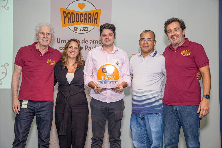 Cinco pessoas posam para a foto em um evento relacionado à padaria. No centro, um homem segura um troféu que diz 'Melhor Padaria de São Paulo'. Ao fundo, há um banner com o logo 'Padocaria SP-2023' e a frase 'Um ato de amor com o pão'. As pessoas estão sorrindo e vestindo camisetas com a marca do evento.