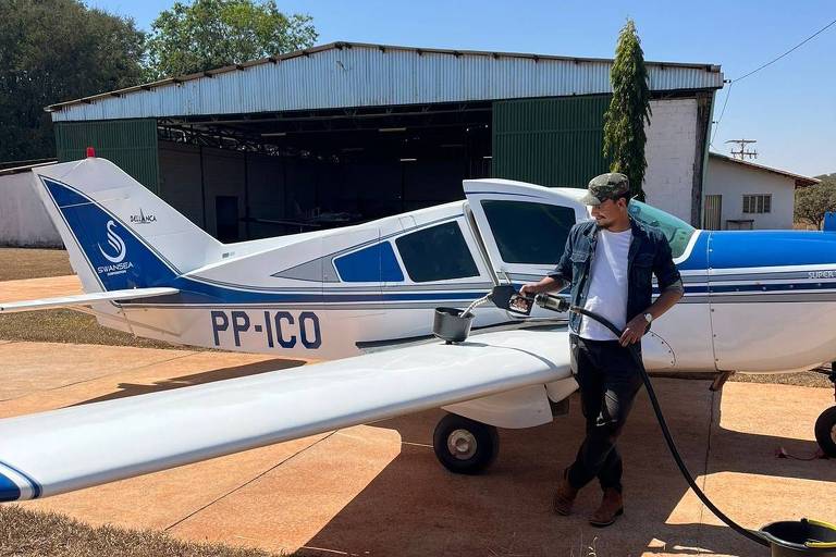 Homem de boné, camiseta e camisa por cima, calça e tênis segura uma mangueira e a acopla em um suporte nas asas do avião. O avião é pequeno, tem cor branca e detalhes azuis, e está pousado em uma espécie de garagem