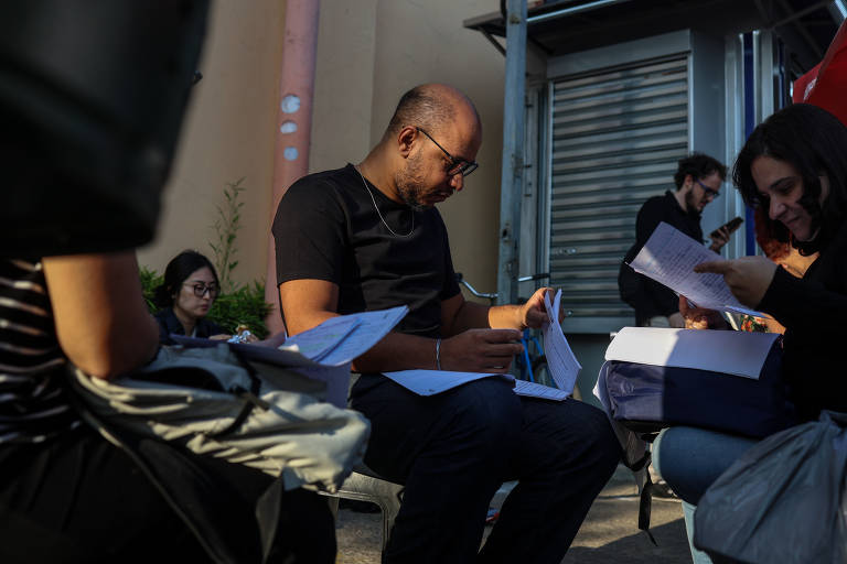 A imagem mostra um grupo de pessoas sentadas em um ambiente urbano. Um homem calvo， usando óculos e uma camiseta preta， está concentrado em papéis que tem em mãos. Ao fundo， outras pessoas também estão envolvidas em atividades semelhantes， com algumas segurando documentos. O cenário parece ser uma área externa， com uma estrutura metálica ao fundo e luz natural iluminando a cena.