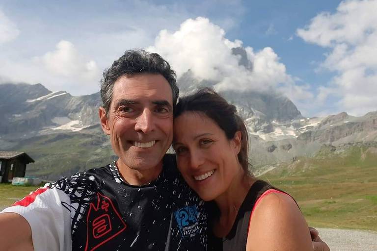 Um casal sorridente tira uma selfie em um ambiente montanhoso. Ao fundo, há montanhas cobertas de nuvens e um céu parcialmente nublado. O homem usa uma camiseta preta com detalhes brancos e vermelhos, enquanto a mulher veste uma blusa preta com detalhes em vermelho. Ambos parecem felizes e estão próximos um do outro.