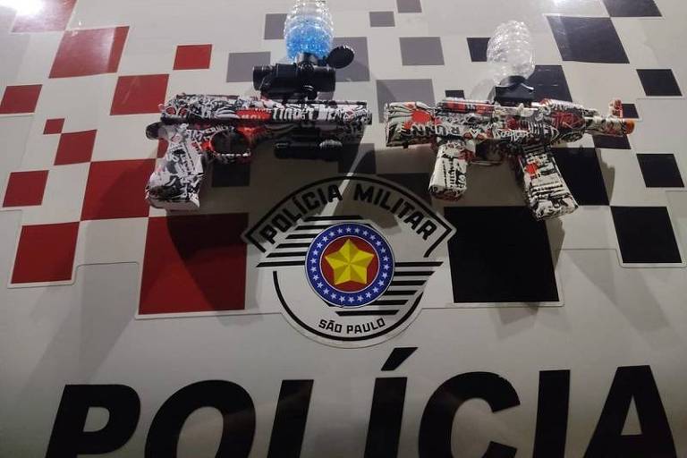 A imagem mostra duas armas de brinquedo, uma em vermelho e outra em preto e branco, sobre o capô de um veículo da Polícia Militar de São Paulo. O fundo é decorado com um padrão de quadrados vermelhos e pretos, e no centro está o emblema da Polícia Militar, que inclui uma estrela amarela 