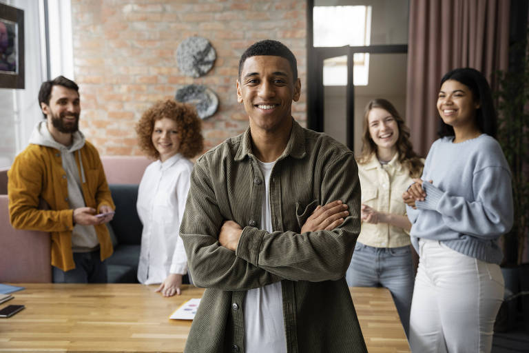 A imagem mostra um grupo de cinco pessoas em um ambiente de trabalho moderno. Um homem de cabelo curto está em primeiro plano, sorrindo e com os braços cruzados. Ao fundo, há uma mulher com cabelo cacheado e uma blusa branca, um homem com barba e uma jaqueta amarela, uma mulher com cabelo liso e uma blusa azul clara, e outra mulher com cabelo liso e uma blusa clara. O ambiente tem paredes de tijolos expostos e uma mesa de madeira com alguns objetos em cima.