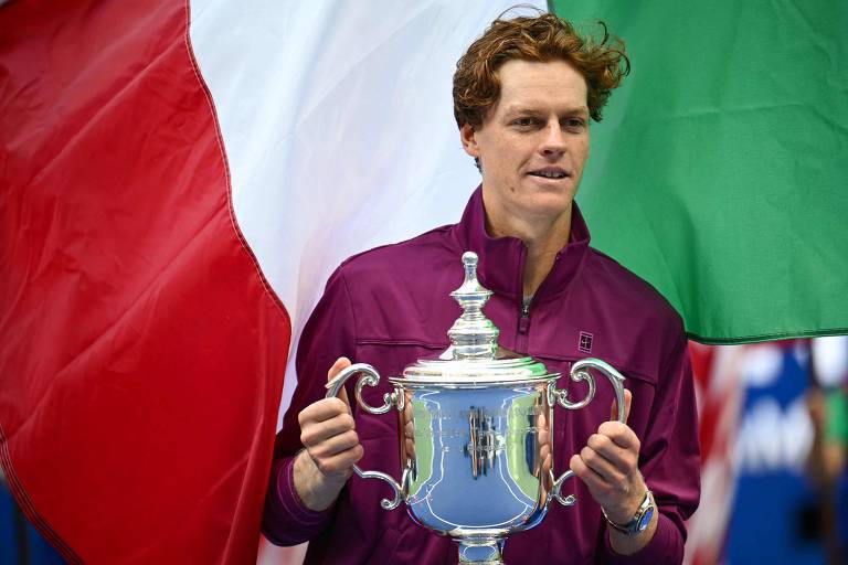 Um jogador de tênis está segurando um troféu em frente a bandeiras. Ele usa uma jaqueta roxa e tem cabelo cacheado. Ao fundo, há uma bandeira vermelha à esquerda e uma bandeira verde à direita.
