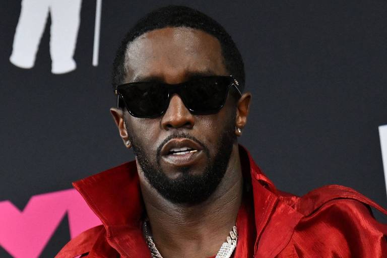 Sean Combs é um homem negro, com óculos escuros, cabelo curto, barba desenhada, que veste um trahe vermelho e uma corrente de ouro no pescoço. Ao fundo, há uma parede preta