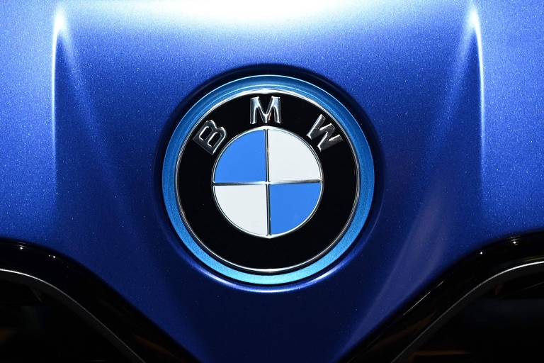 A imagem mostra o logotipo da BMW, que consiste em um círculo dividido em quatro quadrantes, sendo dois azuis e dois brancos, com a sigla 'BMW' em letras prateadas na parte superior. O logotipo está centralizado em um fundo azul metálico brilhante, que apresenta um leve brilho e textura.
