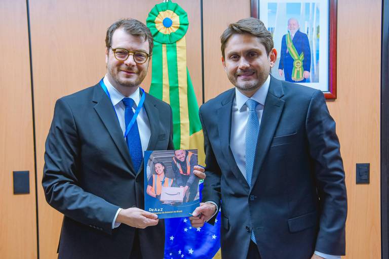 Dois homens em trajes formais estão posando para a foto em um ambiente de escritório. Um deles segura um livro ou documento, enquanto o outro está ao seu lado. Ao fundo, há uma faixa verde e amarela, simbolizando o Brasil, e uma imagem emoldurada na parede.