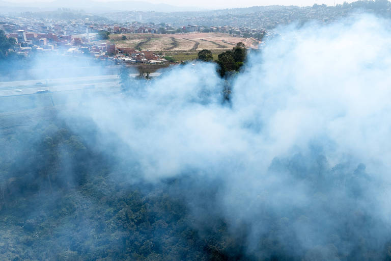 A imagem mostra uma vista aérea de uma área com fumaça densa cobrindo parte da vegetação. Ao fundo, é possível ver uma área urbana com casas e um campo aberto. A fumaça se espalha pelo ar, obscurecendo parcialmente a visão da floresta e da cidade.