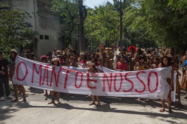 A imagem mostra um grupo de pessoas em uma manifestação, segurando um grande banner branco com a frase 'O MANTO É NOSSO' escrita em letras vermelhas. As pessoas estão vestidas com trajes tradicionais, algumas com penas e adornos na cabeça. O ambiente é urbano, com árvores e edifícios ao fundo, e a luz do sol ilumina a cena.