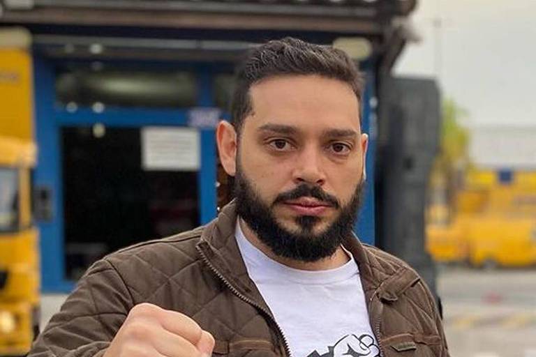 Imagem mostra Ricardo Senese, um homem com barba e cabelo curto, vestindo uma camiseta branca e uma jaqueta marrom, está posando para a foto. Ele levanta a mão em um gesto de punho cerrado, segurando um jornal na outra mão. Ao fundo, há uma construção com paredes amarelas e uma entrada azul.