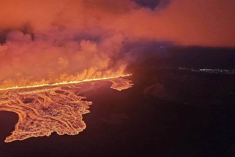 A imagem mostra uma erupção vulcânica com lava fluindo em um padrão sinuoso, cercada por nuvens de fumaça e um céu avermelhado. A lava brilha intensamente, contrastando com a escuridão ao redor, enquanto uma área distante é visível com luzes, possivelmente de uma cidade.