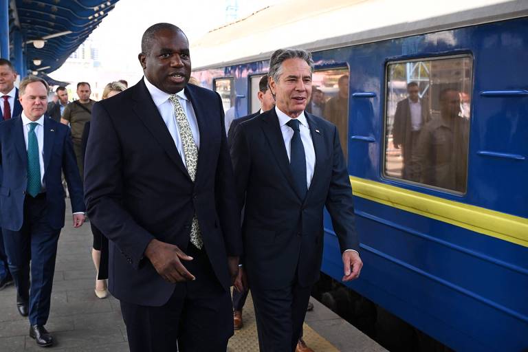 Ao lado de um trem azul com uma faixa amarela estão David Lammy, homem negro de cabelo raspado, e Antony Blinken, homem branco grisalho. Ambos vestem ternos escuros, com Lammy usando uma gravata amarela e Blinken, uma azul.