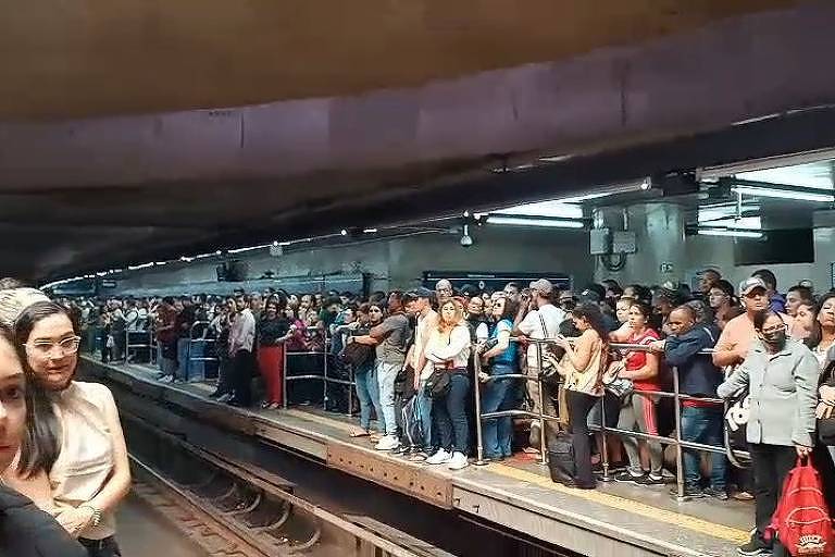 A imagem mostra uma grande multidão de pessoas esperando na plataforma de uma estação de metrô. O ambiente é interno, com iluminação fluorescente e paredes de concreto. As pessoas estão em pé, algumas conversando, enquanto outras olham para o lado. No fundo, é possível ver mais pessoas alinhadas ao longo da plataforma