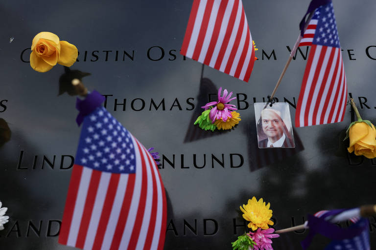 A imagem mostra uma superfície de um memorial com nomes gravados. Pequenas bandeiras dos Estados Unidos estão fixadas na superfície, junto a flores coloridas. Há também uma foto de uma pessoa, que está visivelmente colada ao memorial. O ambiente transmite um sentimento de homenagem e lembrança.