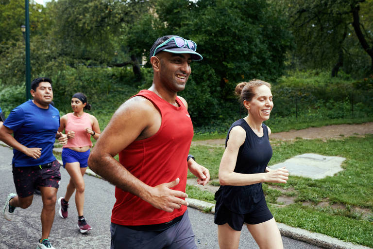 A imagem mostra um grupo de quatro pessoas correndo em um caminho de parque. Um homem à esquerda, vestindo uma camiseta azul e shorts escuros, está correndo ao lado de uma mulher com uma camiseta rosa e shorts. À direita, um homem com uma camiseta vermelha e um boné está sorrindo enquanto corre, acompanhado por uma mulher com uma camiseta preta e shorts. O fundo é composto por árvores e vegetação.
