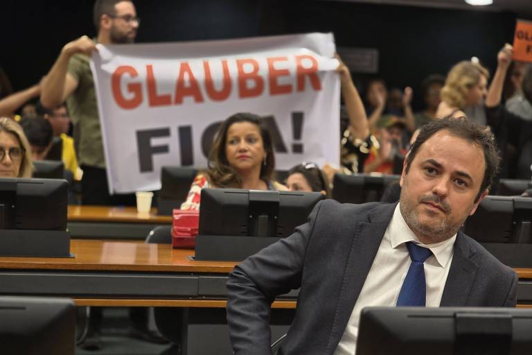 Deputado sentado, de terno e gravata, com apoiadores ao fundo, onde se lê o cartaz "Glauber fica"