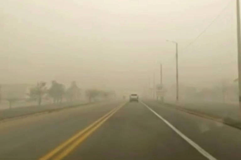 A imagem mostra uma estrada em condições de neblina densa, com visibilidade reduzida. Há um carro branco à frente, e postes de luz estão visíveis ao longo da estrada. O ambiente parece desolado, com árvores e edifícios indistintos ao fundo devido à névoa