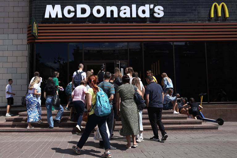 A imagem mostra um grupo de pessoas em frente a um restaurante McDonald's. Algumas pessoas estão subindo as escadas de entrada, enquanto outras estão na calçada. O letreiro do McDonald's é claramente visível acima da entrada.