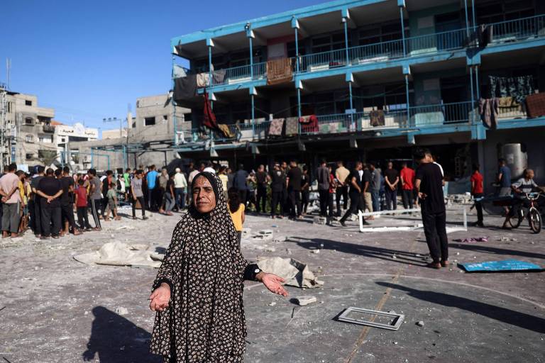 A imagem mostra uma mulher em primeiro plano, vestindo uma roupa escura e com as mãos levantadas, em um cenário de destruição. Ao fundo, há um edifício de vários andares com danos visíveis, e um grupo de pessoas se reúne em meio a escombros e destroços. O céu está claro.